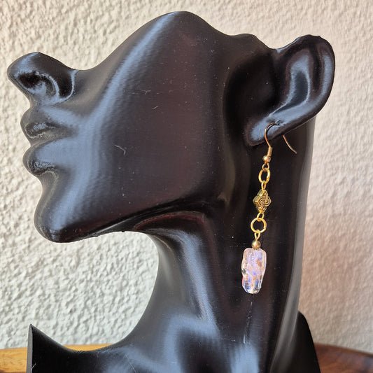 A gold-toned dangle earring with a faceted lavender-purple glass charm, displayed on a black mannequin head against a textured beige wall.