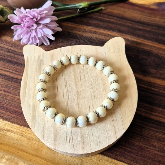 A beaded stretch bracelet with cream-colored round beads and small metallic spacer rings, displayed on a light wood cat-shaped stand over a dark wood surface with a pink flower in the background.